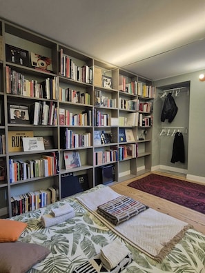 A room with a large bookshelf filled with books, a bed with a patterned bedspread, and a red rug. - 09 - Loft Library - Studio Apartment, Sleeps 2 (Oggebbio)