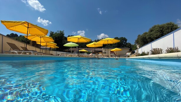 En swimmingpool med kristallklart blått vatten, omgiven av gula och gröna parasoller och solstolar.