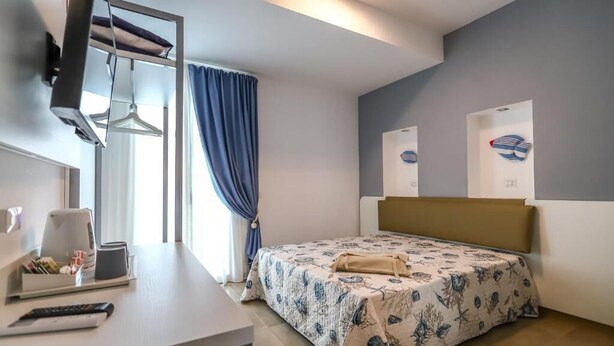 Una habitación de hotel con una cama, un escritorio con silla, un televisor y un ventanal con cortinas.