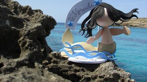 Una figura de una sirena sobre una tabla de surf, con un paisaje de costa rocosa y mar azul cristalino.