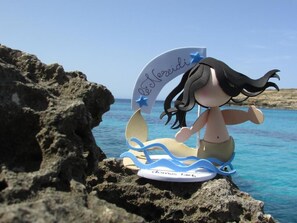 A figurine of a mermaid on a surfboard with a backdrop of a rocky coastline and clear blue sea.