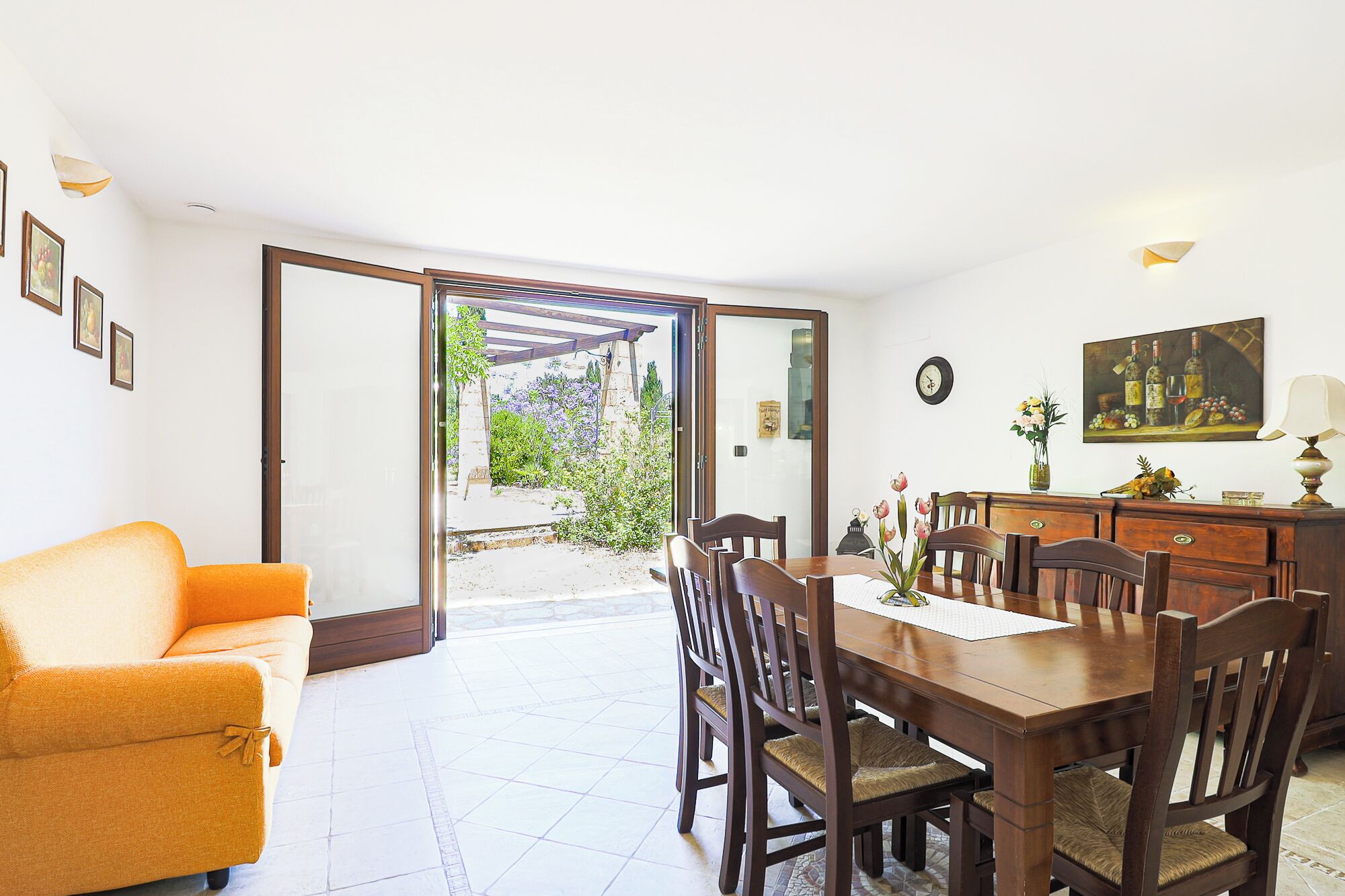 Zona pranzo con tavolo e sedie in legno, un divano e vista su un giardino attraverso una porta aperta.