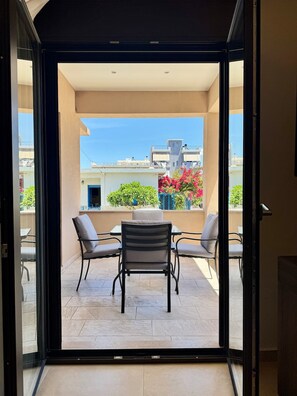 An open sliding glass door leading to a balcony with a table and chairs, a view of buildings, and a clear sky.