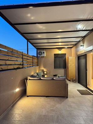 A covered outdoor seating area with a wicker sofa, a table, and a wall-mounted air conditioning unit.
