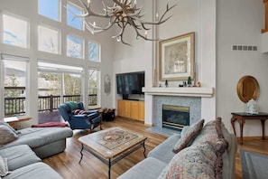 A spacious living room with a fireplace, large windows, a television, and a ceiling chandelier. - NEW! Red Hawk Haven - Sauna, Hot Tub, Game Room (Silverthorne)