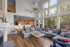 A spacious living area with a large sofa, a coffee table, and a fireplace. There is a kitchen in the background and large windows offering a view of the outdoors.