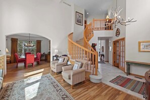 A spacious living area with a staircase, a chandelier, and a dining area with red chairs.