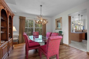 A dining area with a glass table, red upholstered chairs, a chandelier, and a view through large windows.