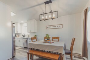 A dining area with a table, chairs, a bench, a hanging light fixture, and a kitchen in the background.