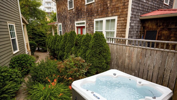 A backyard with a hot tub, surrounded by a wooden fence, dense greenery, and a house with a red door. - Beach Walk, Hot Tub, Pool, Gym, Fire Pit, Games, Sleeps 13 (Lincoln City)