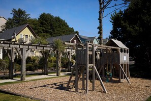 Ein Spielplatz mit Holzgerüsten, darunter eine Schaukel und eine Kletterkonstruktion, umgeben von einem Kiesbereich.