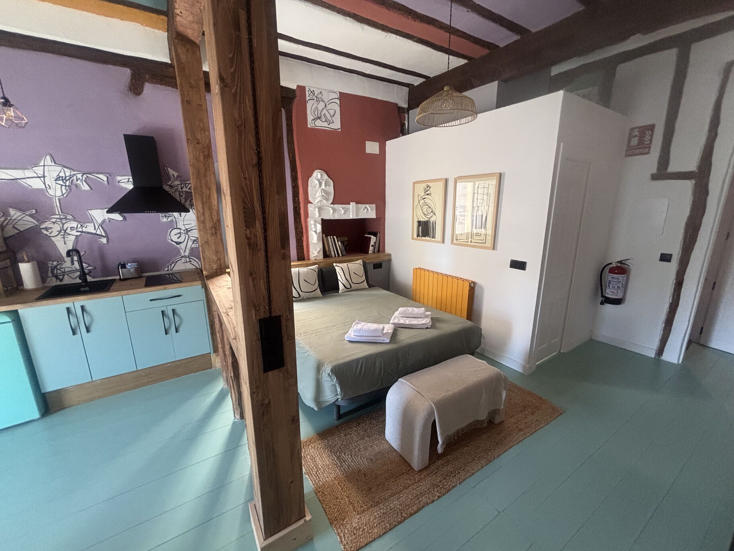 A bedroom with a bed, bedside tables, a wooden beam structure, a radiator, a fire extinguisher, and a wall with artwork.