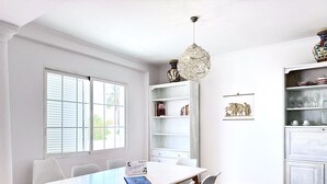 Een moderne keuken met witte kastjes, een eettafel met stoelen, een hanglamp en een raam met jaloezieën.