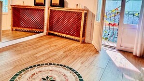 Huoneessa on puulattia, pyöreä mosaiikkikuvioinen matto, puinen konsolipöytä, jossa on koristeellinen ritiläkuvio, sekä liukuovi, joka johtaa ulkoilmassa olevalle parvekkeelle.