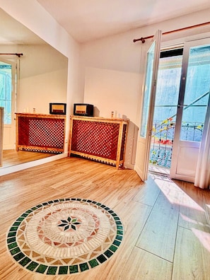 Ein Zimmer mit Holzboden, einem runden Teppich aus Mosaiksteinen, einem Holzkonsolentisch mit Netzmuster und einer Glastür zum Außenbalkon.