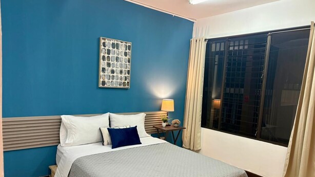 A hotel room with a bed, a nightstand, a lamp, a window with curtains, and a wall art piece.