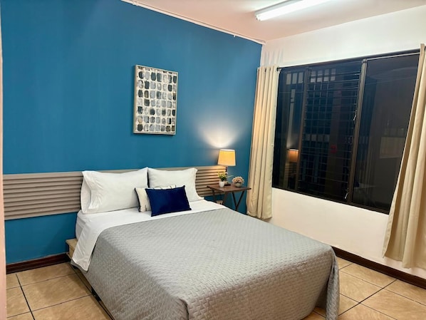 A hotel room with a bed, a nightstand, a lamp, a window with curtains, and a wall art piece.