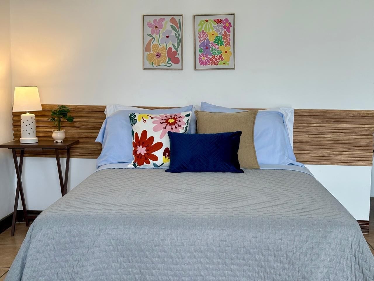 A neatly arranged bed with a grey quilt, a wooden headboard, and two floral wall art pieces.