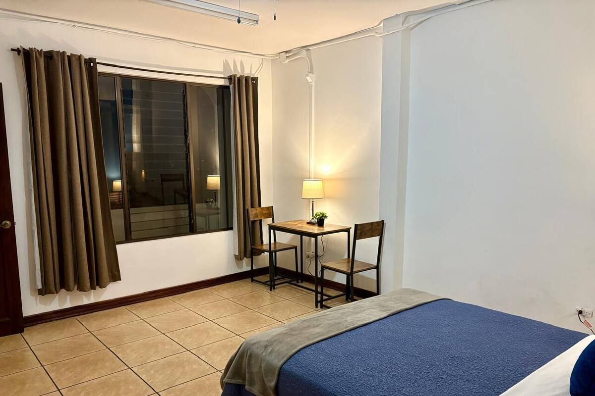 Habitación de hotel con cama, escritorio con sillas, ventana con cortinas y lámpara.