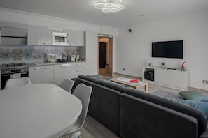 A modern kitchen with white cabinets, a microwave, and a gas stove. There is a dining table with chairs, a sofa, and a TV mounted on the wall.