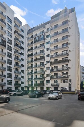 A multi-story residential building with balconies, air conditioning units, and a parking area with cars.