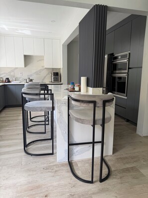 A modern kitchen with a marble countertop, a built-in oven, and a bar area with stools. - Upscale Caribbean Retreat with Oceanview Balcony & Resort Style Living (Montego Bay)