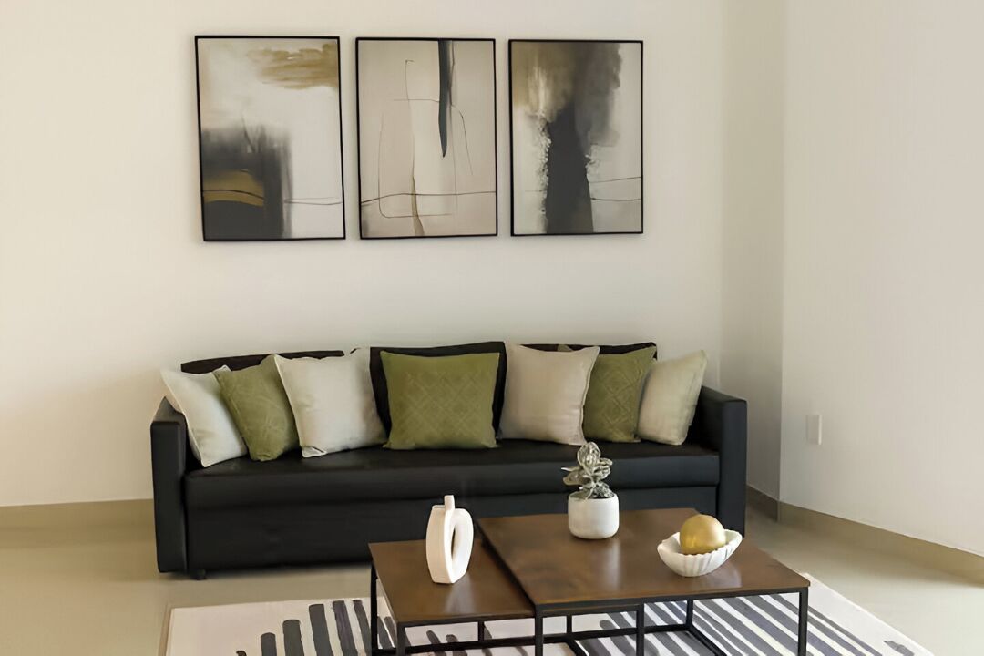 Un salon moderne avec un canapé noir, des coussins verts et blancs, une table basse sur laquelle sont posés un vase blanc et un bol, et trois tableaux abstraits accrochés au mur.