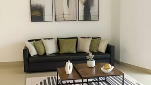 Una sala de estar moderna con un sofá negro, cojines verde y blanco, una mesa de centro con un jarrón blanco y un cuenco, y tres cuadros abstractos en la pared.