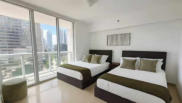 Habitación de hotel moderna con dos camas, un ventanal grande con vistas a la ciudad y un otoman redondito.