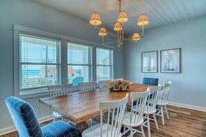 A dining area with a wooden table, white chairs, a chandelier, large windows with blinds, and framed artwork on the wall. - Luxury Stay|Free Linens & Baby Items|Walk to Beach|High End Design and Amenities (Surf City)