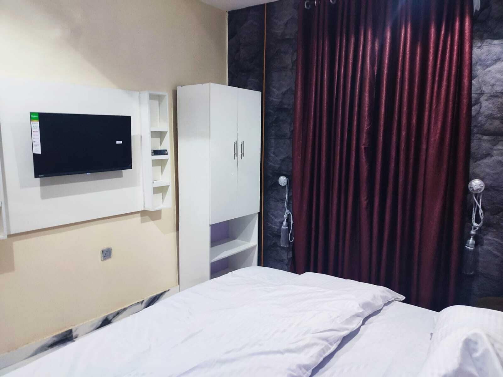 Habitación de hotel con cama, televisión montada en la pared, armario y zona de ducha con cortinas.