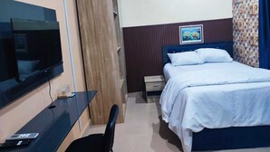 Habitación de hotel con cama, escritorio con televisor, silla, mesita de noche y un cuadro en la pared.