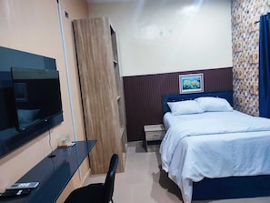 A hotel room with a bed, a desk with a TV, a chair, a nightstand, and a wall-mounted picture. - Regent Hotel and Suites (Osogbo)