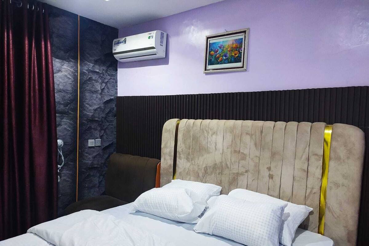 Una habitación de hotel con cama, unidad de aire acondicionado, un cuadro en la pared y una cortina.