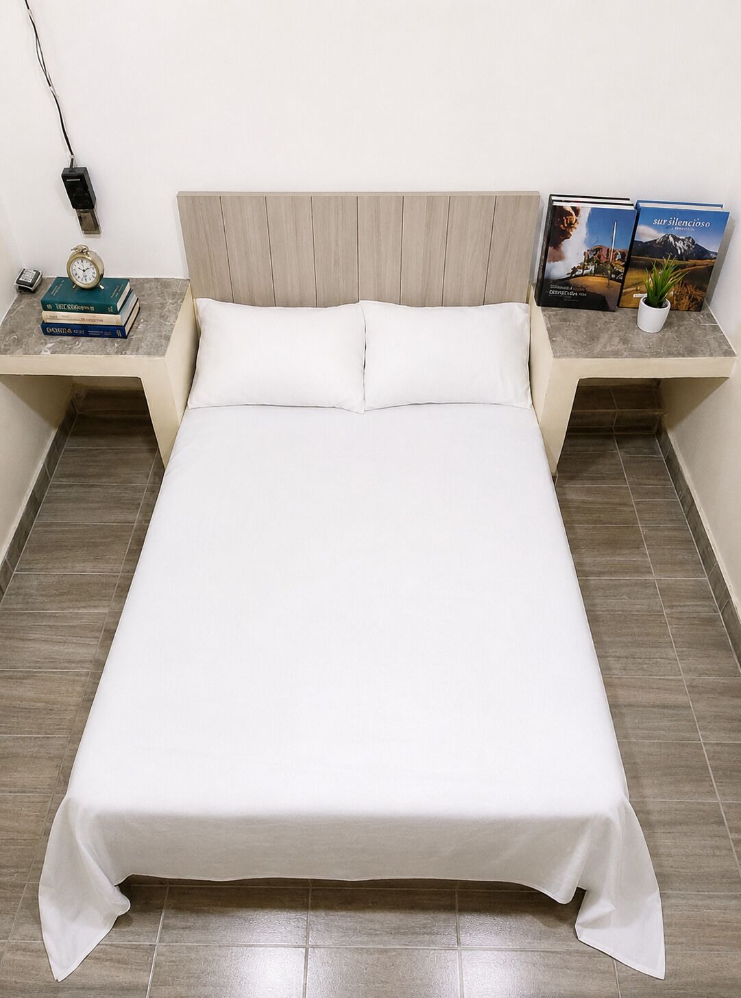 Un lit avec une literie blanche, deux tables de chevet avec des livres et une horloge, et une prise de courant fixée au mur.