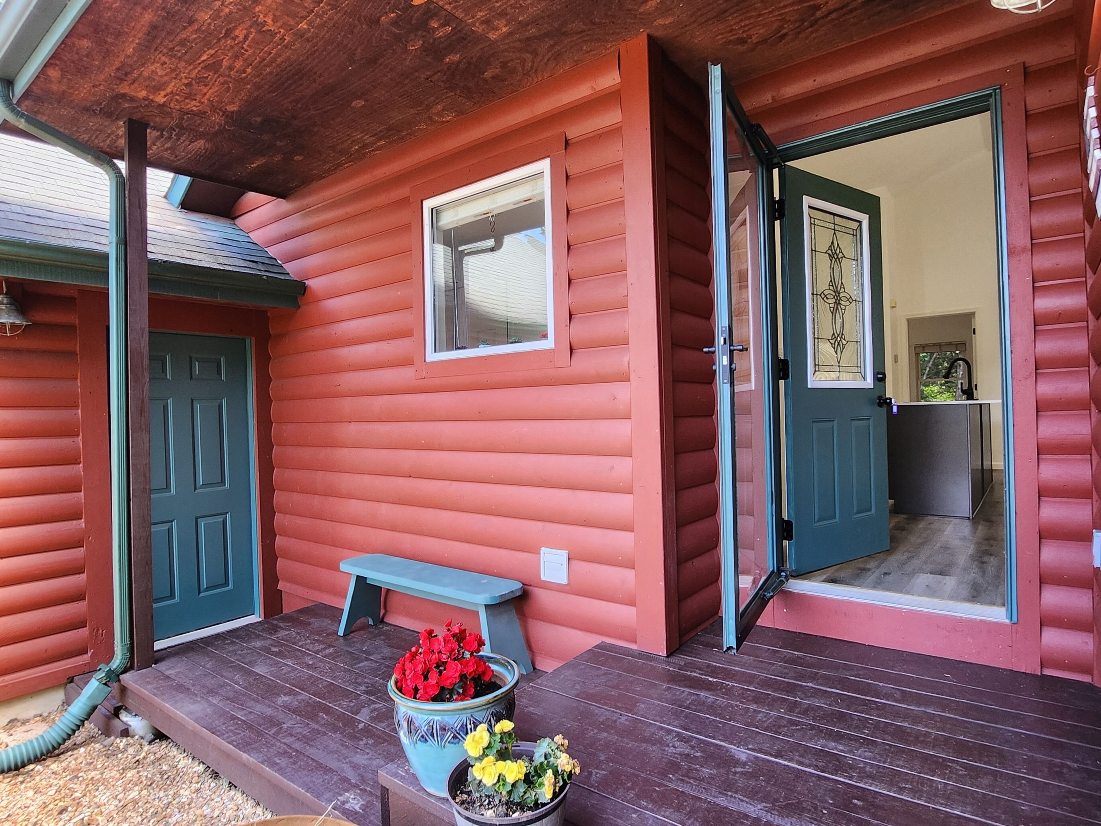 A red wooden building with a porch, a door, a bench, and potted plants.
