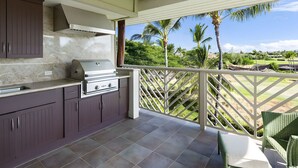 An outdoor kitchen with a grill, adjacent to a balcony with a view of a golf course and palm trees.