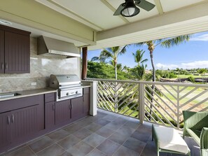 An outdoor kitchen with a grill, adjacent to a balcony with a view of a golf course and palm trees.