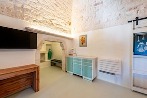 A room with a stone wall, a wooden bench, a teal cabinet, a white radiator, and a flat-screen TV mounted on the wall. - Masseria Ramunno Piccola - Anna Suite by Rentbeat (Ostuni)