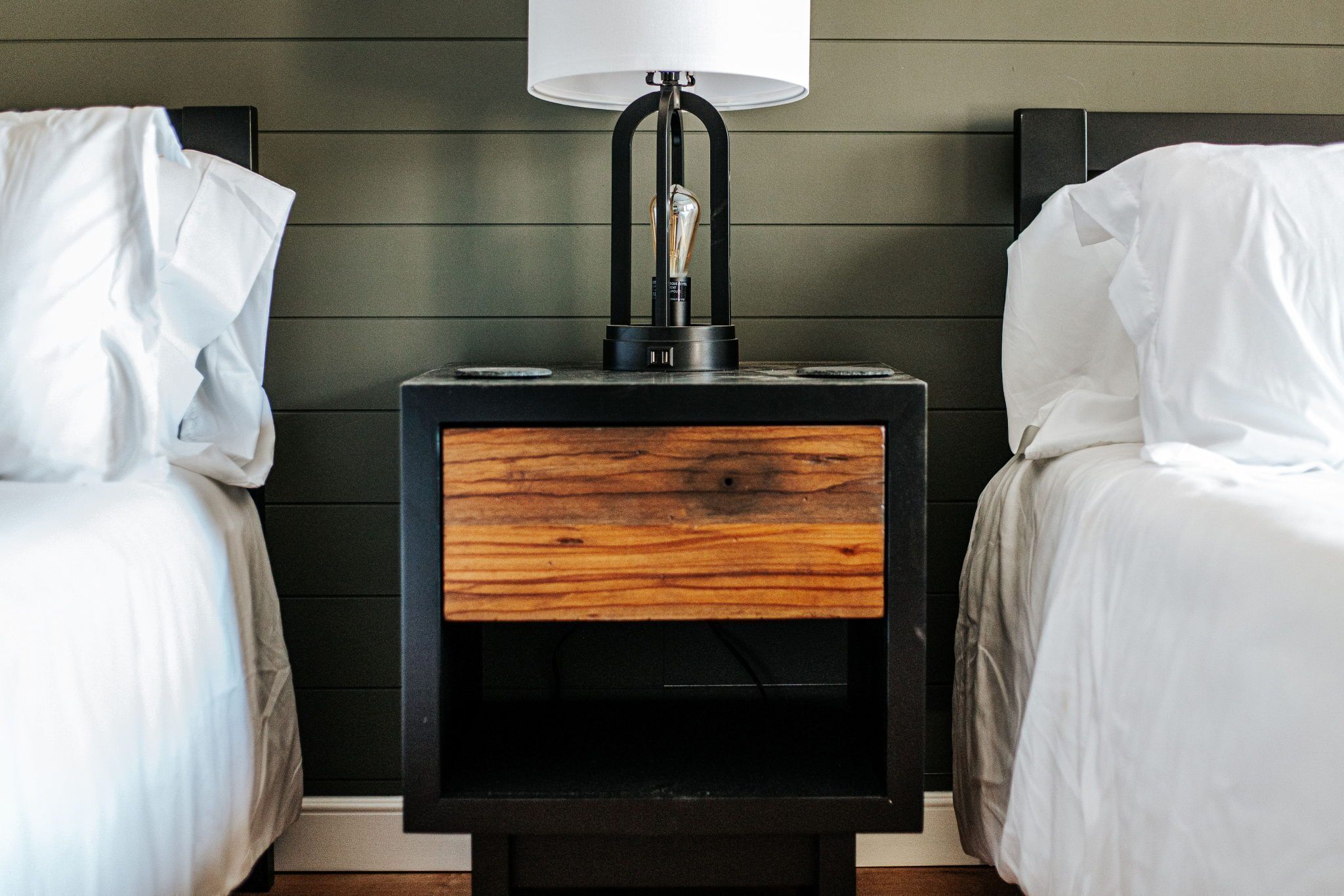A bedroom with two beds, a bedside table with a lamp, and a wooden drawer.