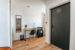 Quarto compacto com piso de madeira, uma escrivaninha com cadeira, um pequeno refrigerador preto, micro-ondas, uma planta em vaso, um espelho e uma porta preta.