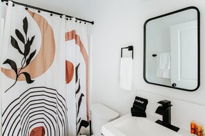 A bathroom with a shower curtain featuring leaf and stripe patterns, a toilet, a sink with a black faucet, and a mirror with a black frame. - Skyline Drive Adventure Base (Waynesboro)