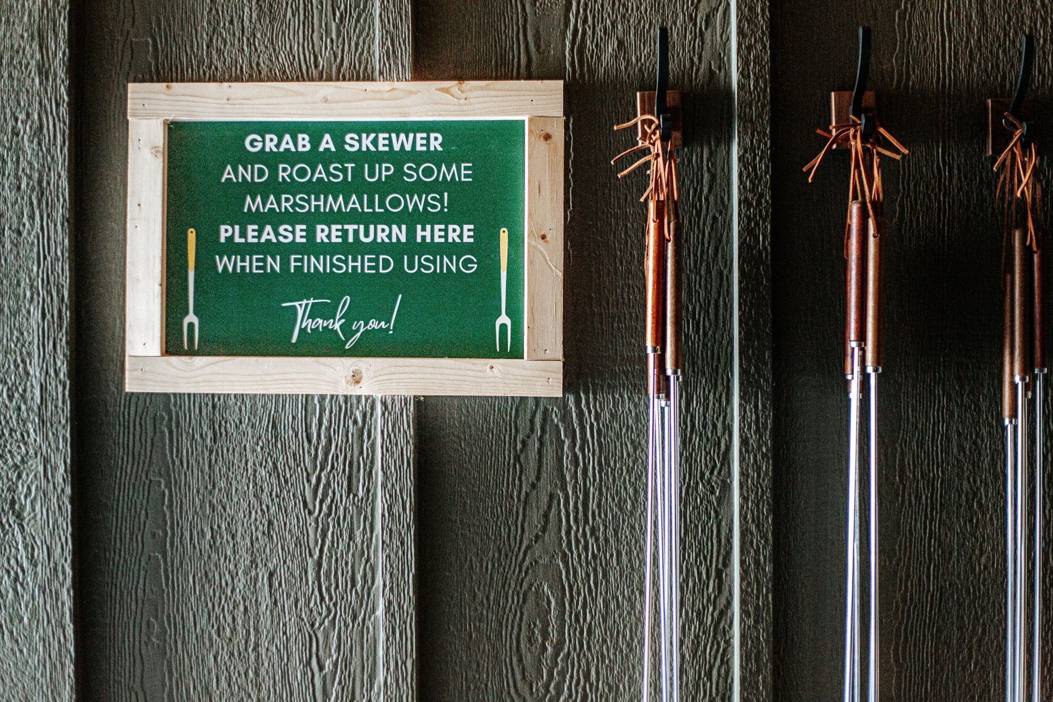 A sign with a skewer and marshmallows, and three skewers on a wooden surface.