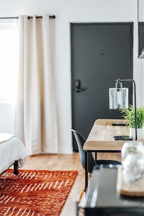 Ein modernes Hotelzimmer mit Bett, Schreibtisch mit Lampe, Topfpflanze und einem Teppich mit rotem Muster.