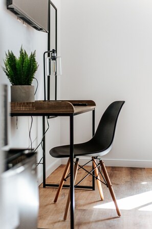 Canto de um quarto com piso de madeira, uma cadeira preta, uma mesa de madeira, uma planta em vaso e um dispositivo fixado na parede.