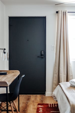Una habitación con una puerta oscura, una mesa de madera con sillas, una cama y un tapete rojo y blanco, y cortinas color beige.
