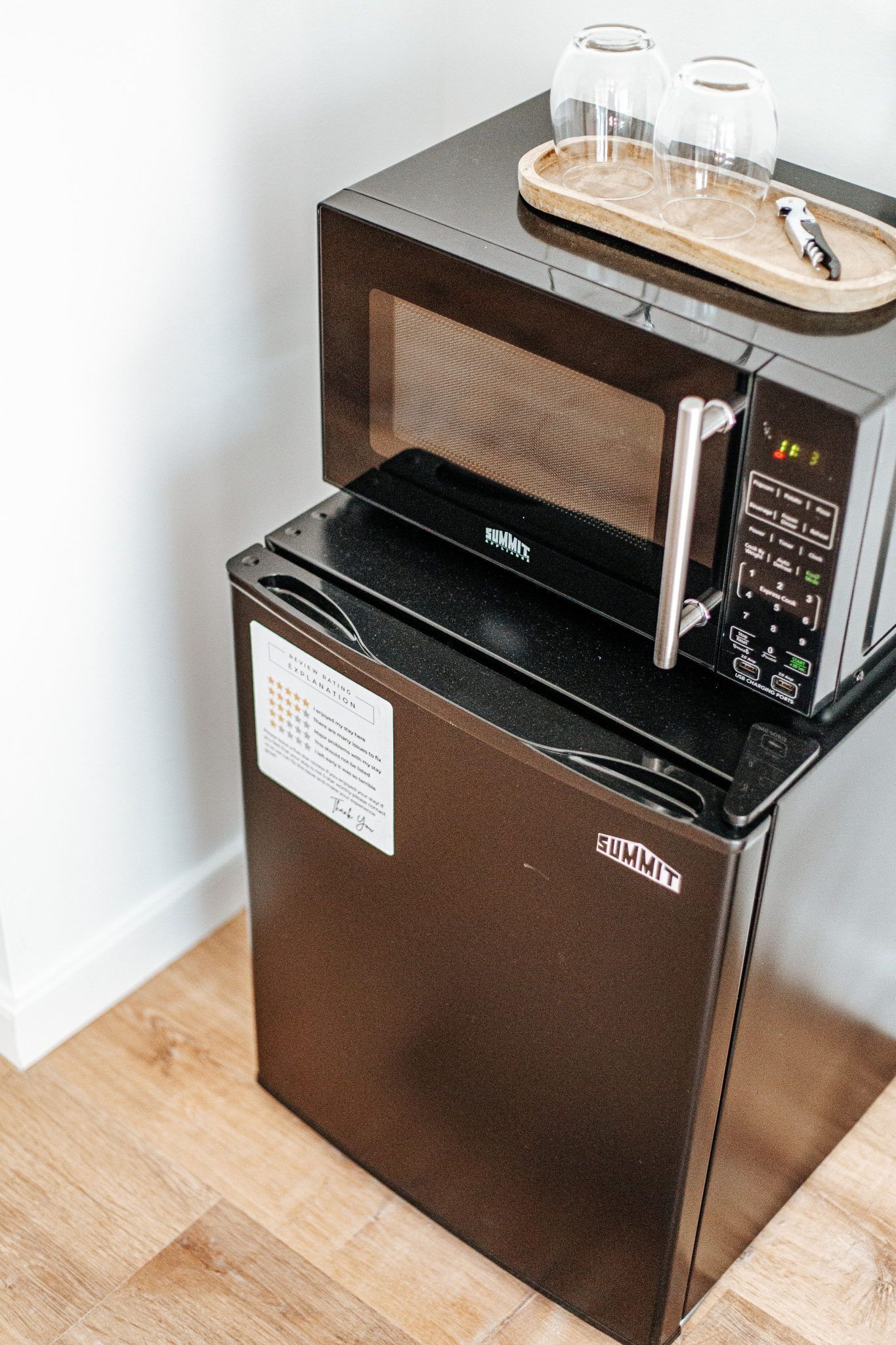 Una cocina compacta con microondas y refrigerador, una bandeja de madera con dos vasos, y un electrodoméstico de acero inoxidable.