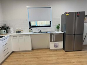 A modern kitchen with white cabinetry, a stainless steel refrigerator, a built-in dishwasher, and a window with blinds. - 3BR Haven in Stony Rise (Stony Rise)