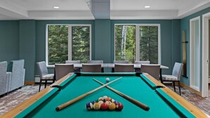 A pool table with balls and cues, surrounded by chairs and tables, with large windows and a view of trees. - Serene location, spaceous with pool and easy access to Banff and Lake Louise (Canmore)