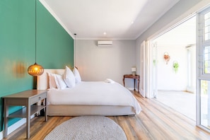 A bedroom with a bed, bedside tables, a hanging light fixture, an air conditioning unit, and a view through a glass door.